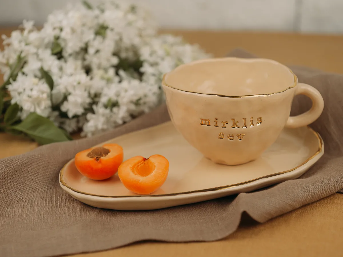 A beige ceramic cup with gold lettering sits on a matching saucer beside two halved apricots, with white flowers in the background, in a soft still life composition.