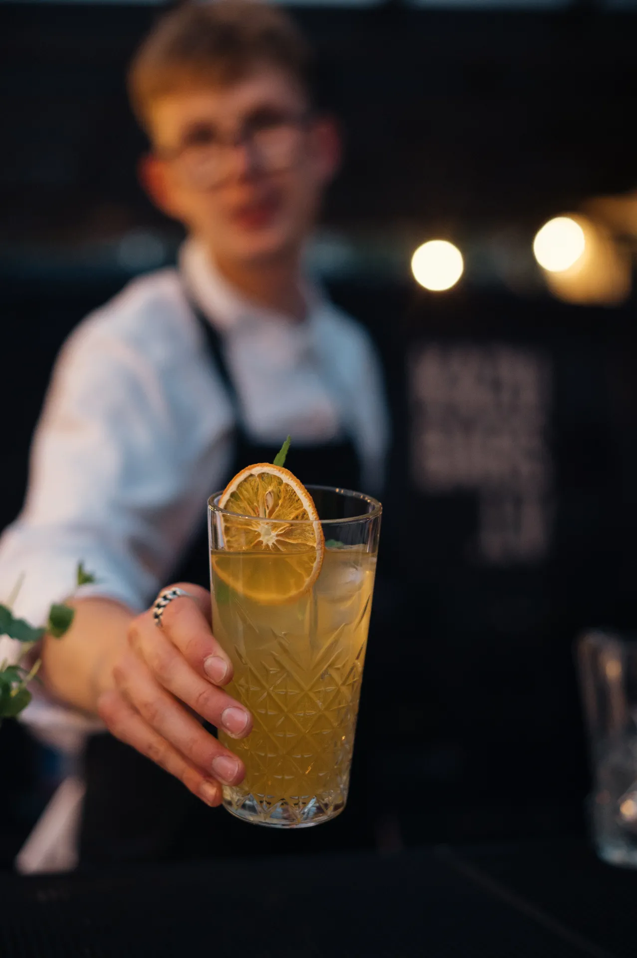 A bartender in a white shirt and black apron extends a patterned highball glass with a yellow cocktail garnished with a dried orange slice toward the camera. The background is softly blurred with warm bokeh lights, creating a shallow depth of field commercial bar scene with a welcoming, intimate mood.