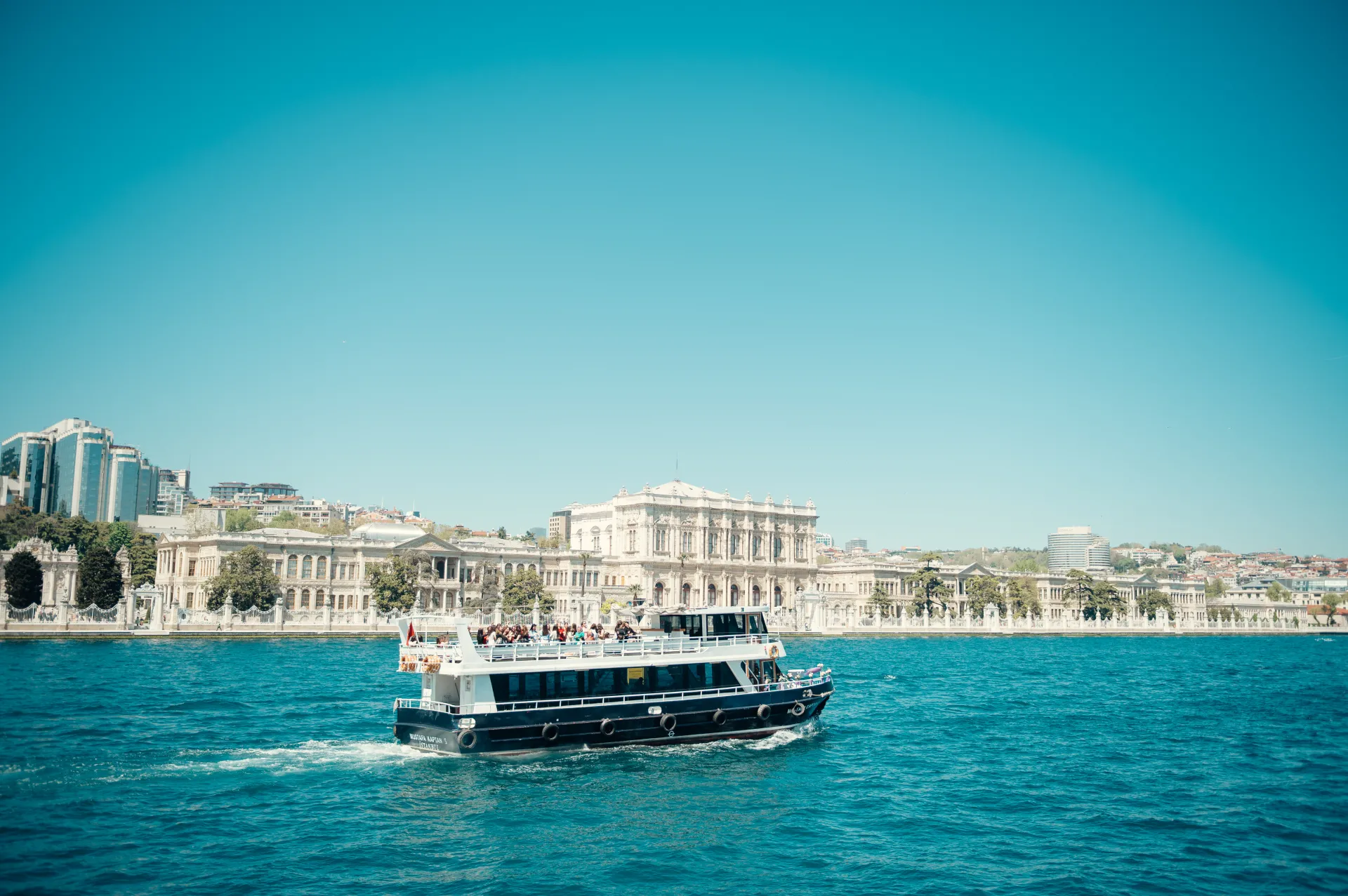 Stambula/Kapadokija — A white and dark blue passenger ferry moves across bright turquoise water, leaving a wake behind it. In the background, a grand classical building stands along the shoreline under a clear blue sky, with modern high-rises visible to the left.