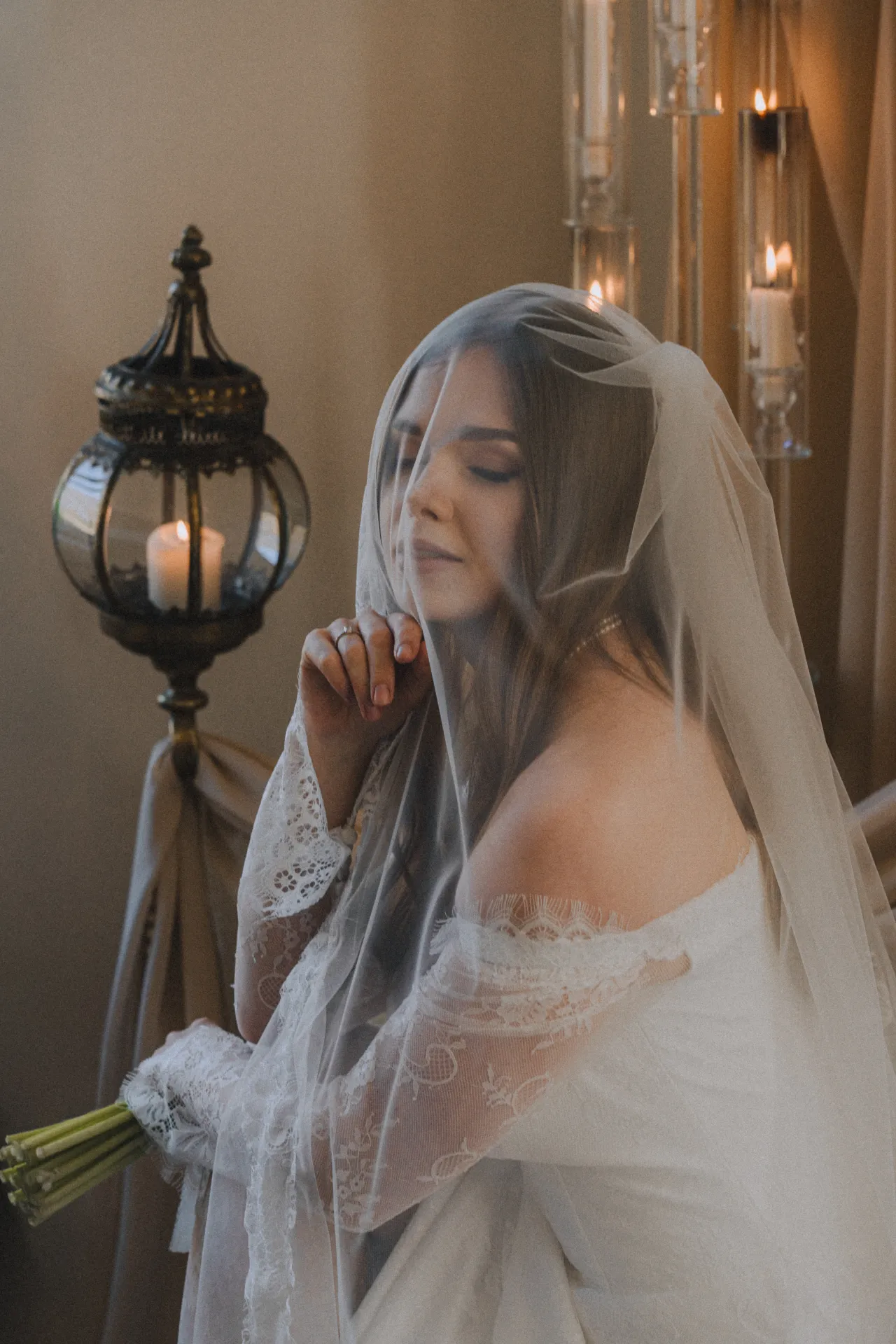 A bride in a white off-the-shoulder lace wedding dress with a sheer veil over her head sits indoors, eyes closed with a hand near her face and a bouquet in her lap. Warm candlelight from an ornate lantern and tall glass candle holders creates a soft, intimate mood in a wedding portrait style.