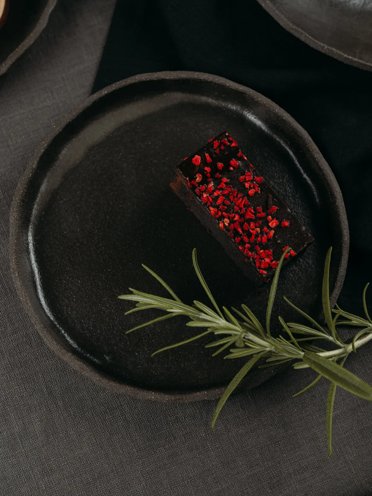 Top-down still life of a dark chocolate brownie topped with red sprinkles on a matte black ceramic plate, with a fresh rosemary sprig resting beside it. The moody, minimalist food styling is set on dark linen, emphasizing texture and contrast in a commercial product photography style.