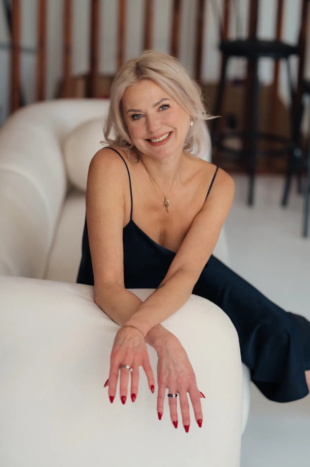 A smiling blonde woman sits on a white rounded sofa in a bright modern interior, wearing a black spaghetti-strap slip dress and posing with her arms crossed on the sofa arm. She has red manicured nails, rings, a delicate necklace and bracelet; the softly blurred background shows wooden slats and black bar stools.
