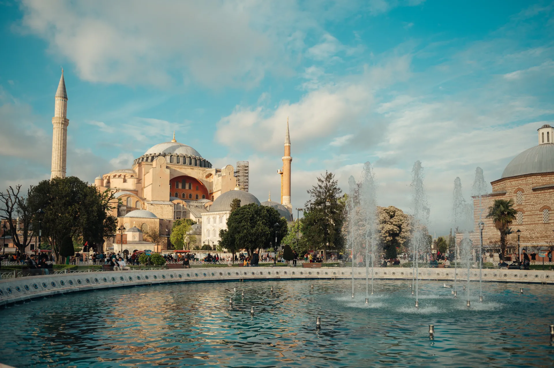 Stambula/Kapadokija — A large historic domed building with two slender minarets sits behind a curved fountain basin with active water jets. Green trees, a domed stone structure on the right, and small groups of people gather along a paved promenade under a bright blue sky with soft white clouds, gi…