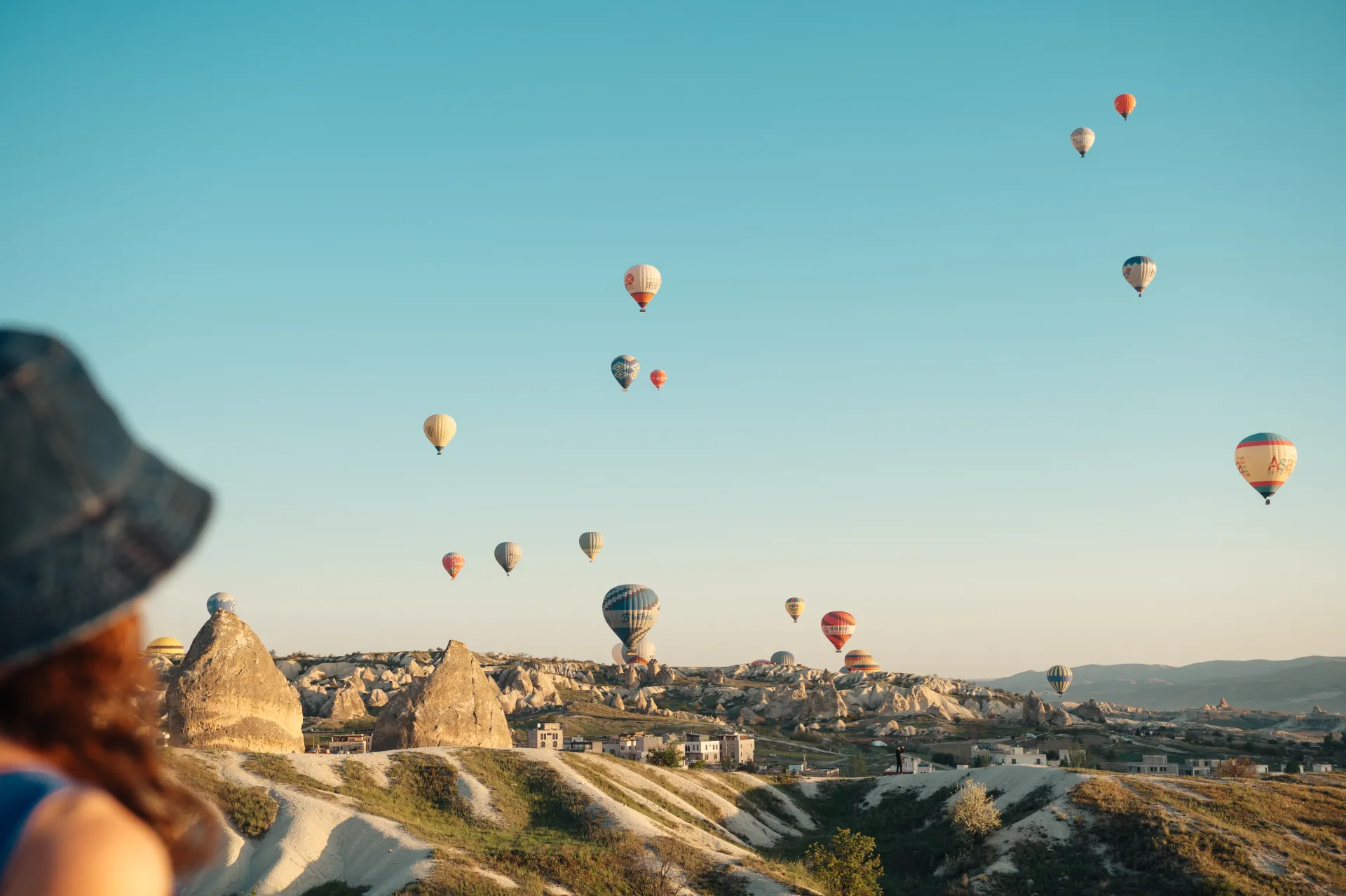 Stambula/Kapadokija — A person wearing a hat watches numerous colorful hot air balloons floating above a rocky, eroded landscape with scattered buildings under a clear sky.