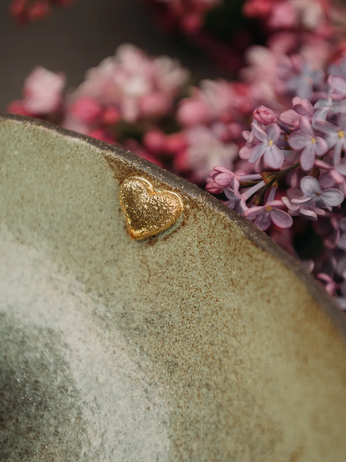 A close-up still life shows a textured ceramic bowl rim with a small gold heart accent, set against a soft-focus background of pink and purple lilac blooms under gentle natural light.