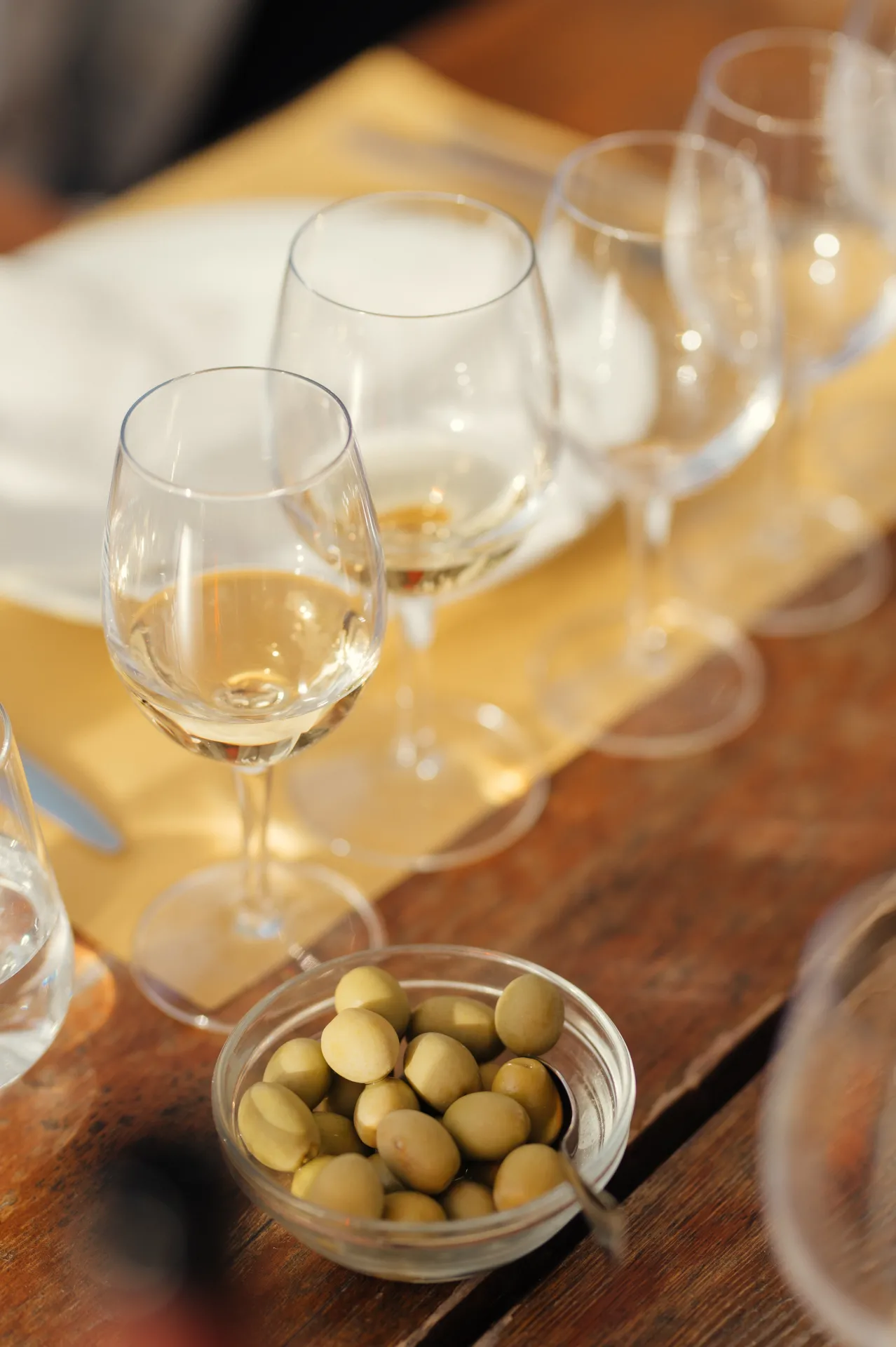 Itālija/Toskāna — A close-up food photography shot shows several wine glasses with white wine arranged on a wooden table with yellow placemats, alongside a small glass bowl of green olives, creating a warm, intimate dining atmosphere.
