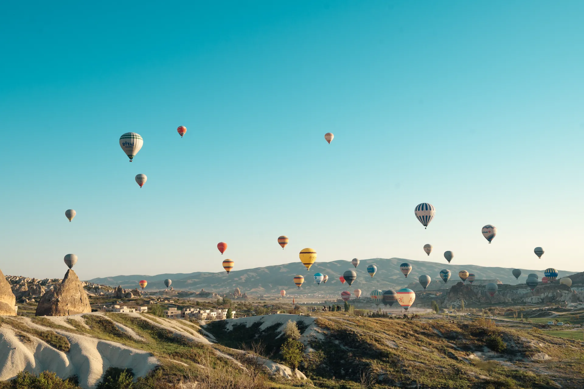Stambula/Kapadokija — Numerous colorful hot air balloons float over a rugged, grassy valley with pale rock formations and distant mountains under a clear turquoise sky. The wide landscape scene captures the balloons at various altitudes in soft morning light, conveying a serene aerial tourism atmos…