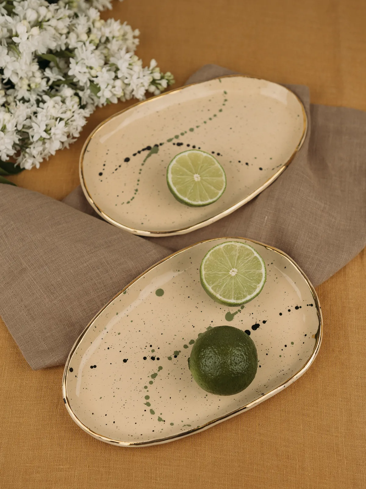 Two oval cream ceramic plates with gold rims and speckled glaze rest on a beige cloth beside white flowers, each holding lime slices and a whole lime in soft natural light, styled as product photography.