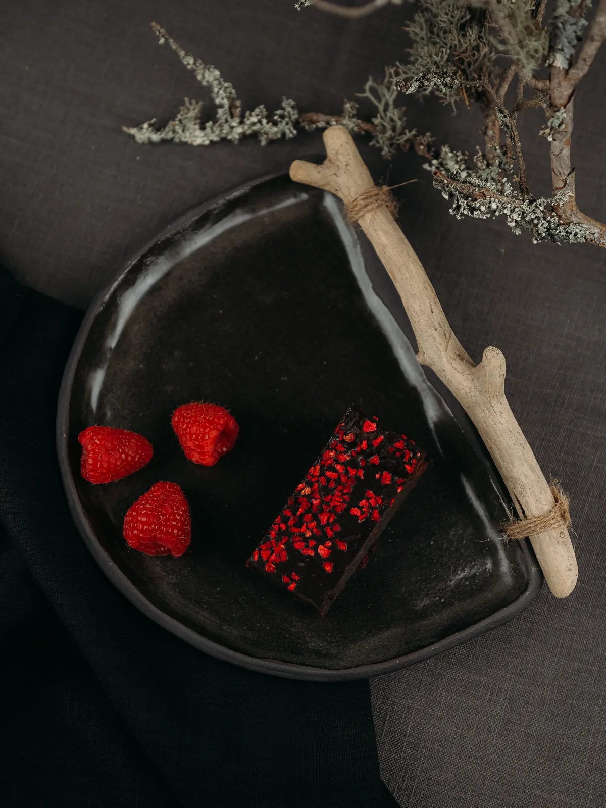 A dark ceramic plate holds three red raspberries and a chocolate bar topped with red sprinkles, accompanied by a light driftwood branch tied with twine. The moody food photography features soft lighting against a dark fabric background with dried branches.