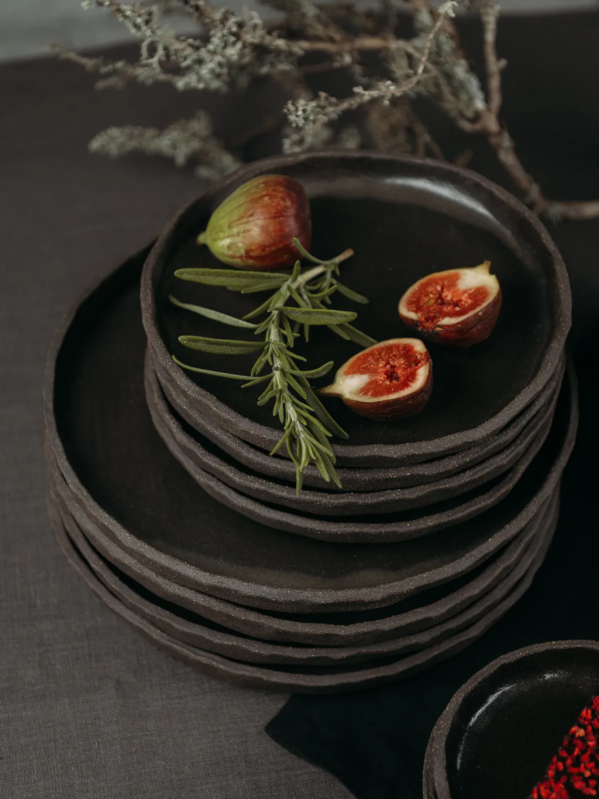 A stack of dark handmade ceramic plates sits on a dark linen surface, topped with a whole fig, two halved figs and a sprig of rosemary. Dried lichen branches and a small dark bowl with red seeds appear in the background in a moody still life product style.