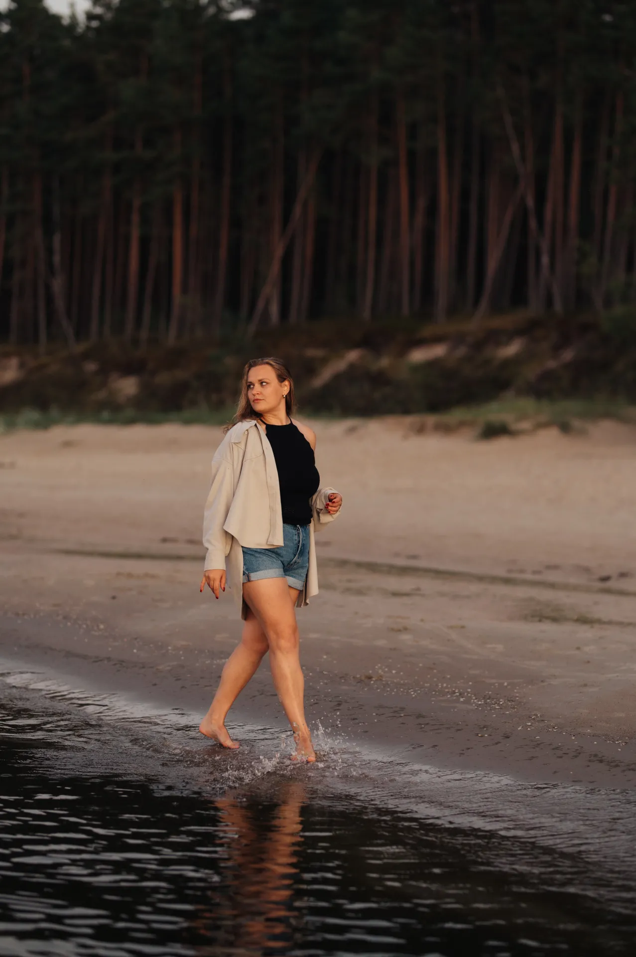 A woman walks barefoot through shallow water at the water’s edge on a sandy beach, wearing a black top, denim shorts and an unbuttoned beige overshirt. Behind her stretches a calm shoreline with dark trees under soft golden-hour light, creating a contemplative outdoor lifestyle portrait.