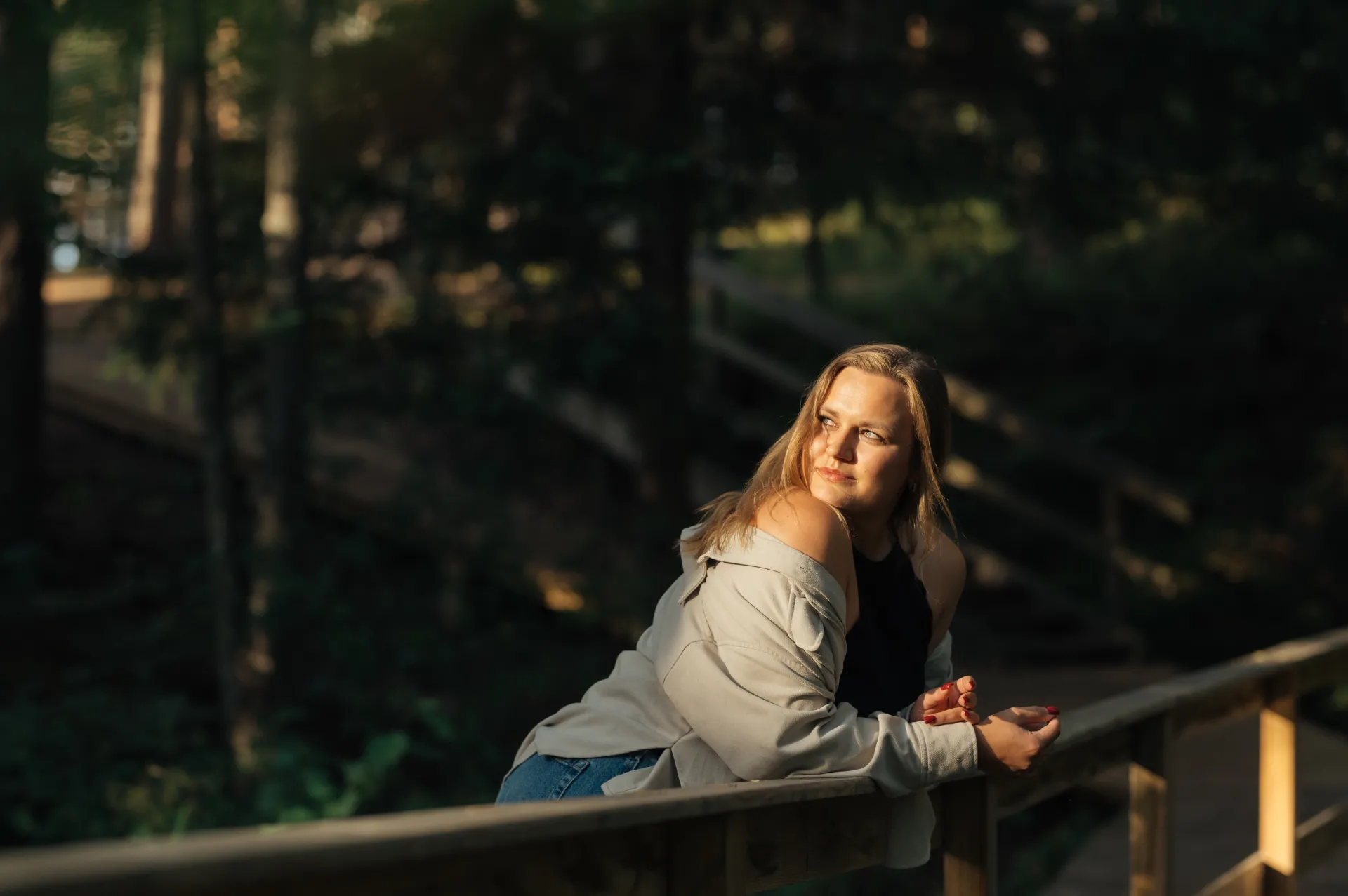 A woman leans on a wooden railing in a sun-dappled forest, wearing an off-the-shoulder light jacket over a black top and blue jeans, looking toward the camera in a natural portrait style.