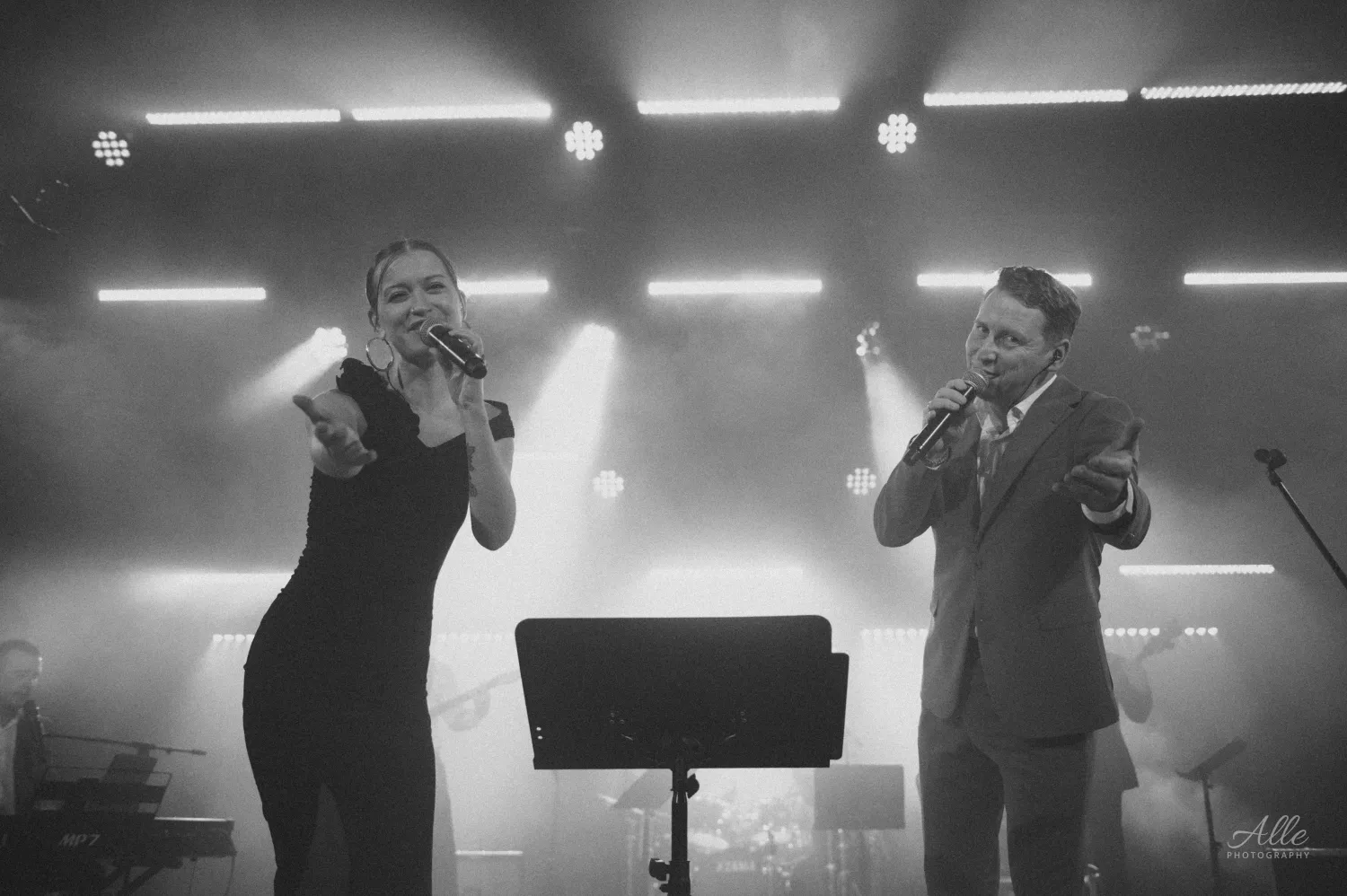 A man and a woman sing into microphones on a smoky stage illuminated by bright overhead lights, with a music stand between them and band equipment visible in the background.