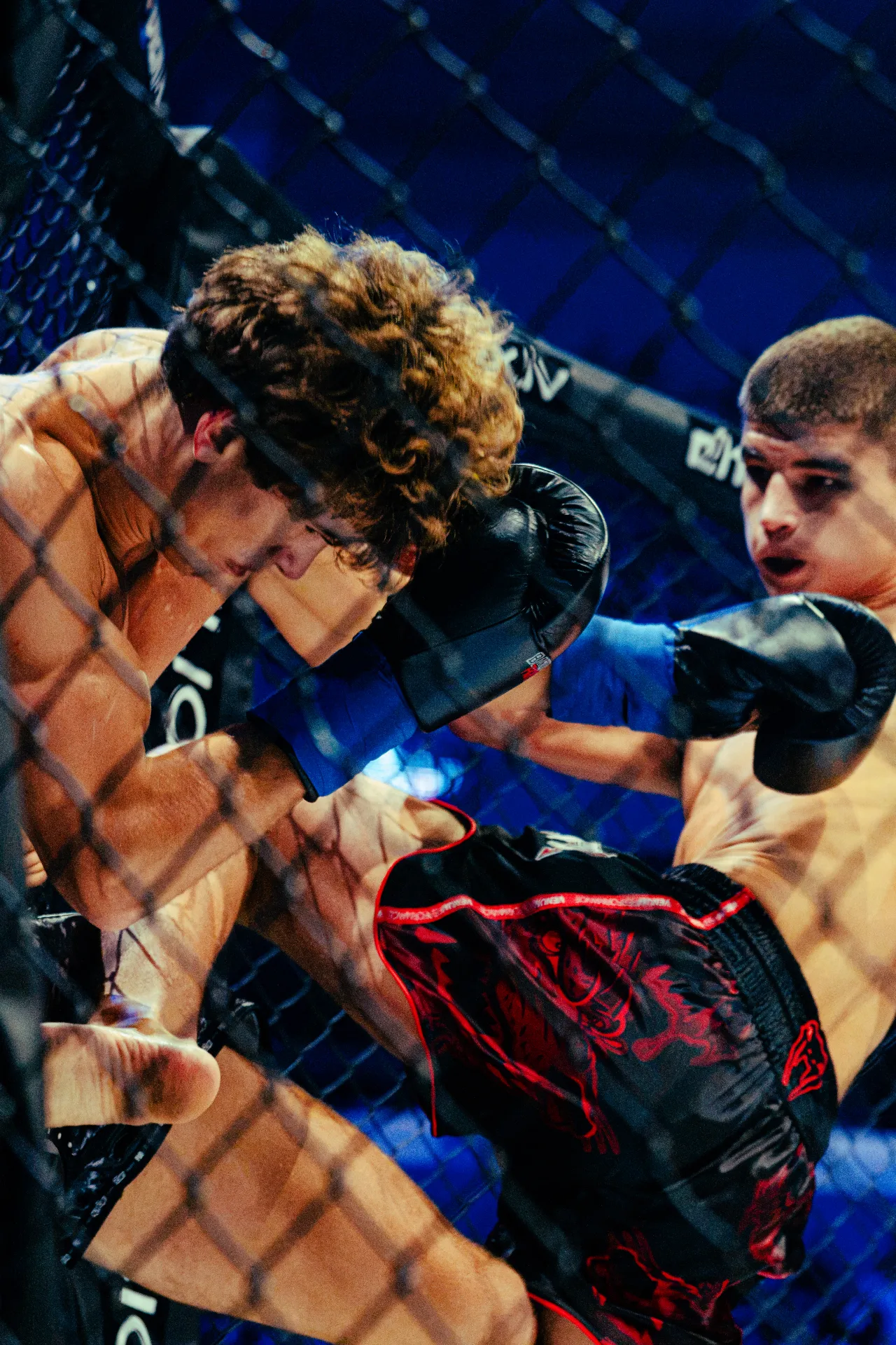 Divi kailkrūtaini cīnītāji ar boksa cimdiem cīnās MMA ringā ar metāla sietu, dramatiskā zilā apgaismojumā. Viens cīnītājs ar cirtainiem matiem atspiežas pret žogu, bet otrs, kurš valkā melnus šortus ar sarkaniem rakstiem, šķiet, ka izdara sitienu ar dūri vai kāju dinamiskā sporta ainas kadrā.