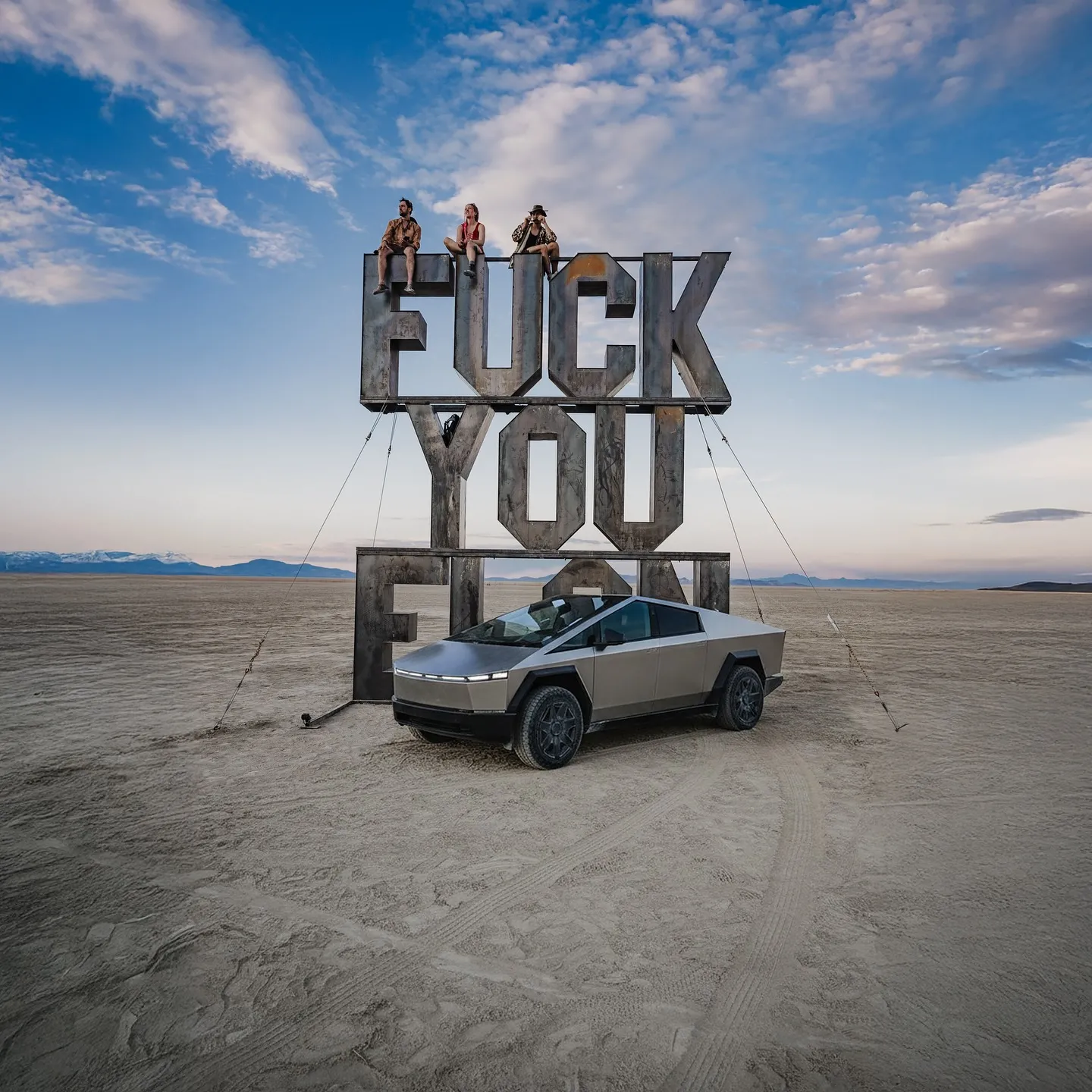 Fuck you Elon — Trīs cilvēki sēž uz lielas metāla skulptūras, kuras uzraksts ir „FUCK YOU”, līdzenā tuksnesī zem apmākušām debesīm, un priekšā ir novietots sudrabkrāsas, leņķains elektromobilis; komerciāla automobiļu fotogrāfija.