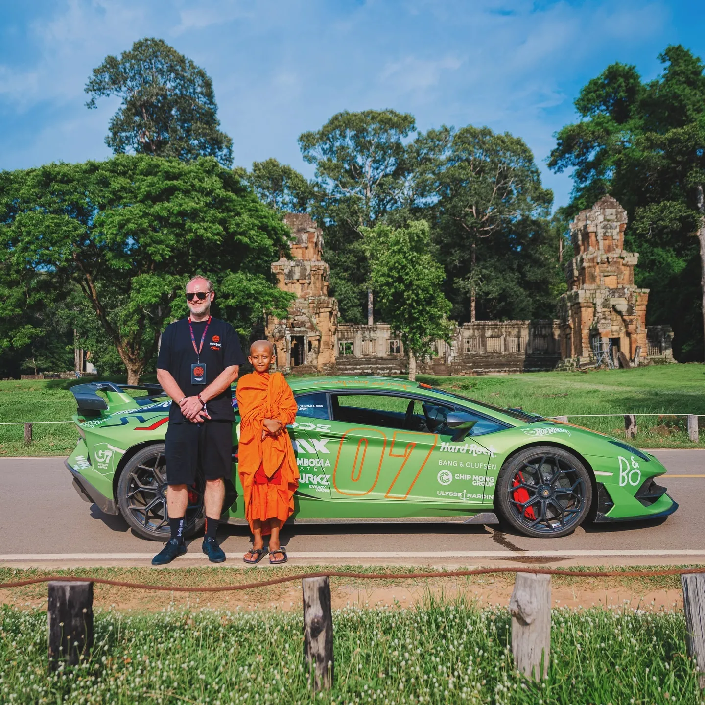 Gumball 3000 — A man in black shirt and shorts stands beside a young monk in an orange robe in front of a bright green sports car with large “07” graphics, on a paved road with grass and wooden posts in the foreground.